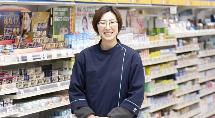 インタビューキービジュアル。Hさんが正面を向いて笑顔で立っている様子。背後には薬の棚がある。