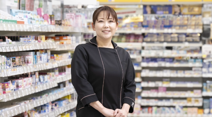 インタビューキービジュアル。Aさんが正面を向いて笑顔で立っている様子。背後には目薬などの商品棚がある。