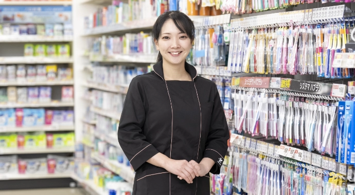 インタビューキービジュアル。Nさんが正面を向いて笑顔で立っている様子。背後には歯ブラシ等の日用品の棚がある。