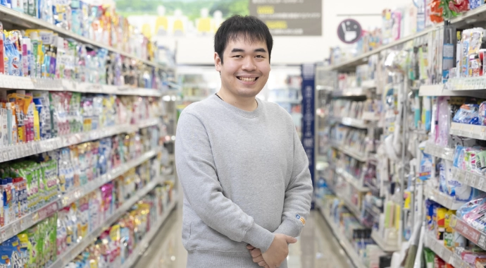 インタビューキービジュアル。Sさんが正面を向いて笑顔で立っている様子。背後には洗濯洗剤などの日用品の棚がある。