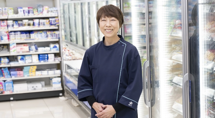 インタビューキービジュアル。Fさんが正面を向いて笑顔で立っている様子。背後には冷凍食品の棚がある。